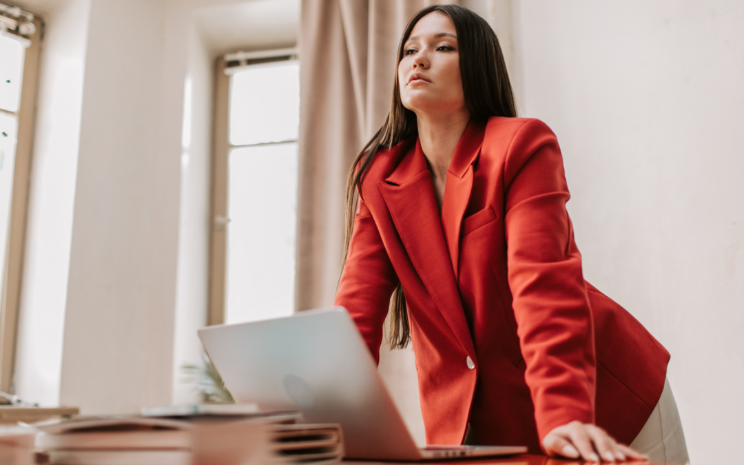 Una donna indossa un blazer rosso, è chinata in diezione della scrivania e sta guardando il suo computer portatile