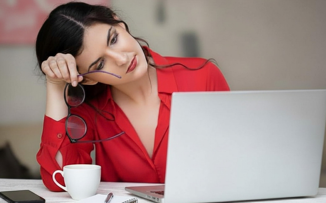 Una donna in camicia rossa, esausta, appoggia la testa sulla mano e guarda un computer portatile, tenendo gli occhiali. simboleggiando la desensibilizzazione alla comunicazione d'urto.